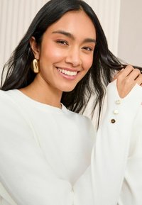 Weißer Pullover mit gerippten Bündchen, verziert mit drei dekorativen Knöpfen in unterschiedlichen Farbtönen: klar, hellbraun und dunkelbraun. Weiche Textur.