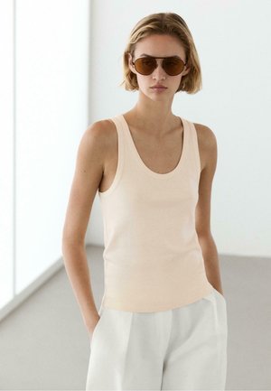 Kvinde med kort blondt hår iført brune solbriller, beige ærmeløs top og hvide bukser, står med hænderne i lommerne i et lyst rum.