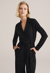 Melnā saplacināta kombinezona ar garām piedurknēm, strukturētu audumu, apkakles kakla izgriezumu un sasietu jostu. Izmanto sānu kabatas. Vienkāršs, elegants dizains.