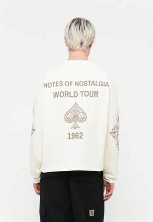 Long-sleeve cream sweatshirt with brown text reading "NOTES OF NOSTALGIA WORLD TOUR 1962" and floral designs on sleeves.