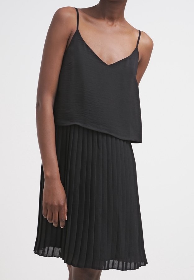 Femme portant un haut noir sans manches à col en V avec fines bretelles et une jupe plissée noire taille haute sur fond uni.