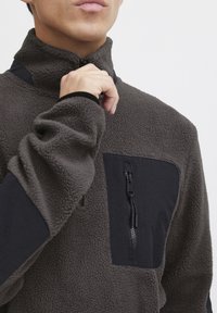 Fleece genser i mørkegrå, med høy krage, svarte tekstildetaljer, brystlomme med glidelås og teksturert, mykt materiale.