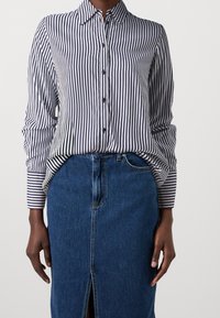 Camicia a righe blu navy e bianca, con maniche lunghe e colletto a punta, abbinata a una gonna di jeans con spacco anteriore.