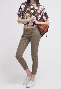 Floral cropped top with short sleeves, olive green fitted pants, and white sneakers. Tan backpack with a shoulder strap.