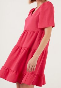 Růžové šaty s n ěkolika vrstvami, V-výstřihem a krátkými nabíranými rukávy, vyrobené z lehké tkaniny s texturovaným povrchem a volánkovým lemem.