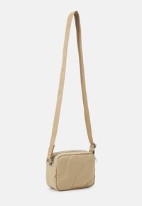 Beżowa pikowana torba na ramię z zapięciem na zamek i teksturowanym paskiem z powtarzającym się wzorem logo. Kompaktowy prostokątny kształt.
