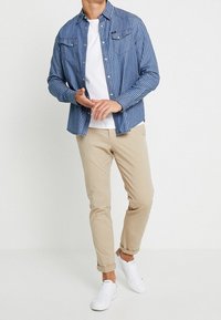 Gestreept blauwe casual blouse over een wit T-shirt, kaki broek en witte sneakers. Voorzien van knoopsluiting aan de voorkant en accenten op de borstzak.