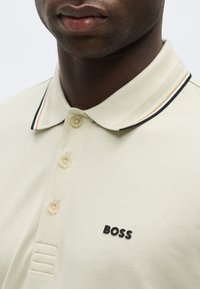 Κοντινό ενός ατόμου που φοράει ένα μπεζ πόλο πουκάμισο με ριγωτό γιακά και μαύρο λογότυπο "BOSS" στο στήθος.