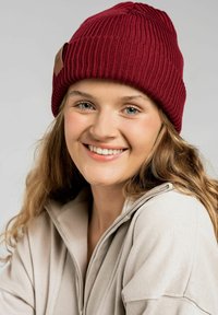 Czerwony, prążkowany beanie z dzianiny z skórzaną metką, noszony na długich, falowanych włosach. Modelka ma na sobie beżową bluzę na zamek. Tło jest w neutralnym odcieniu szarości.