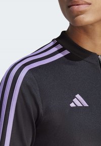 Giacca da track nera con zip, con strisce viola e nere sulle spalle; presenta una texture morbida e un piccolo logo Adidas bianco sul lato sinistro.