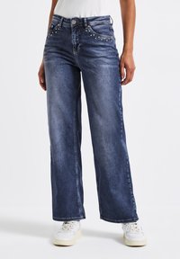 Jean en denim à jambes larges de couleur bleu foncé, avec des clous décoratifs le long des poches avant et un design classique à cinq poches.