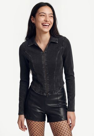 Jeune femme souriante, portant un haut noir zippé à manches longues, un short en cuir noir et des collants résille, sur fond clair uni.