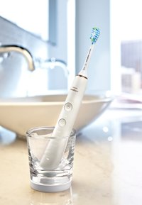 Spazzolino elettrico con manico bianco, setole blu e verdi. È poggiato in un bicchiere trasparente su un piano in marmo, con un lavandino sullo sfondo.