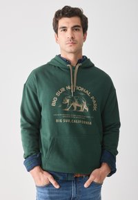 Sweatshirt à capuche vert foncé avec un graphique d'ours à l'avant et du texte. Il possède des poignets côtelés et une poche kangourou à l'avant. Porté par-dessus une chemise à carreaux.