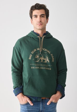 Donker groene hoodie met een vooraanstaande grafiek van een beer en tekst. Bevat ribgebreide manchetten en een voorste kangaroo-pocket. Gestyled over een geruit shirt.