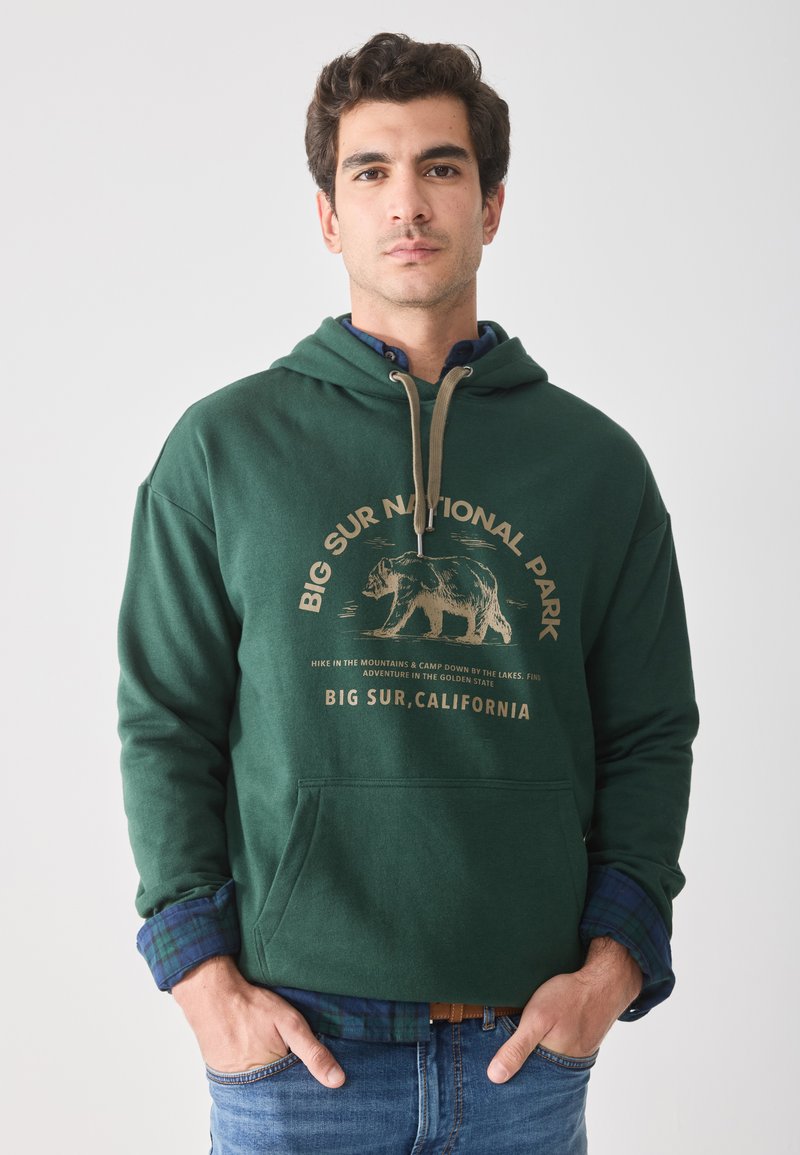 Sweatshirt à capuche vert foncé avec un graphique d'ours à l'avant et du texte. Il possède des poignets côtelés et une poche kangourou à l'avant. Porté par-dessus une chemise à carreaux.