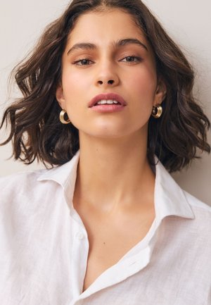White linen shirt with a collar and button details. Model has wavy brown hair and gold hoop earrings, with a natural makeup look.