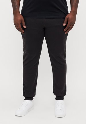 Homme portant un pantalon de jogging noir resserré aux chevilles et des baskets blanches, debout devant un fond clair uni.