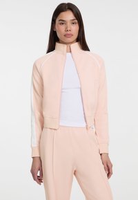 lichtroze zip-up jack met hoge kraag, witte zijstrepen, zachte textuur en aansluitende manchetten, gecombineerd met bijpassende broek.