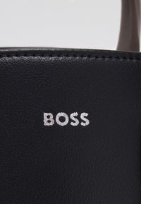 Czarny skórzany materiał o delikatnej fakturze; wyróżnia się srebrnym logo "BOSS"; czyste szwy wzdłuż krawędzi. Prosty, funkcjonalny design.