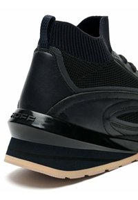 Zapatilla deportiva negra con talón de cuero liso, parte superior de malla y suela texturizada con detalles claros y parte trasera elevada para mayor soporte.