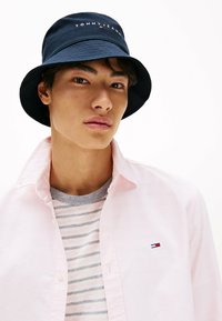 Marineblauwe bucket hat met het "TOMMY JEANS" logo, gedragen met een lichtroze overhemd over een grijs gestreept T-shirt.