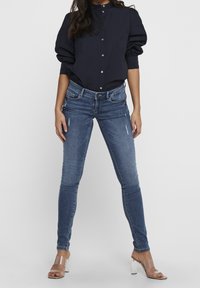 Mujer vestida con una blusa azul marino abotonada, jeans ajustados rasgados de color azul y sandalias de tacón transparente, de pie con las piernas abiertas contra un fondo blanco.