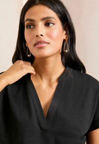 Melna V-veida kakla blūze ar īsām piedurknēm, gluda tekstūra un brīva piegriezuma. Raksturīga smalka spīduma un minimāla šuvju detaļa.