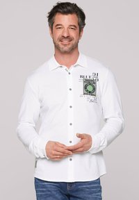 Camicia bianca a maniche lunghe con chiusura a bottoni, caratterizzata da testo grafico e un logo circolare sul petto. Materiale in cotone, vestibilità comoda.