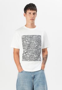 Hvit bomullst-skjorte med en svart grafisk trykk med ulike uttrykksfulle doodles og tekst i et kvadratisk design. Ledig passform, kort erm.