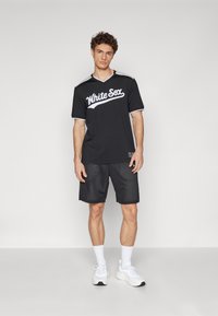 Crna sportska majica s tekstom "White Sox", kratkim rukavima i bijelim detaljima, u kombinaciji s crnim mrežastim hlačama i bijelim tenisicama.