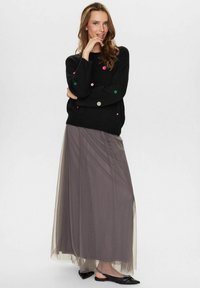 Pull noir avec des pois colorés, associé à une longue jupe grise transparente. Le mannequin se tient dans une pose décontractée avec des ballerines noires.