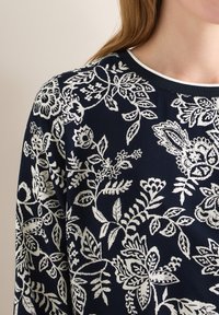 Tissu bleu marine avec un motif floral et feuillu blanc, porté par une personne à peau claire et aux longs cheveux bruns, montrant le cou et l'épaule.