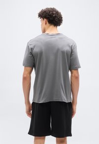 Άνδρας με σγουρά μαλλιά που φοράει απλό γκρι μπλουζάκι και μαύρο σορτς, στέκεται με την πλάτη προς το μέρος μας μπροστά σε λευκό φόντο.