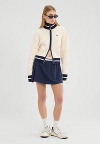 Ciężki sweter zapinany na zamek w kolorze kremowym z granatowymi akcentami, w połączeniu z granatową plisowaną spódnicą w stylu tenisowym. Całość dopełniają białe buty sportowe.