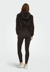 M�ørkebrunt, fluffy fleece hoodie med hætte, parret med sorte ribbede leggings og lyse sportssko.