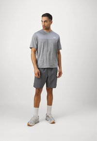 Grå atletisk t-skjorte med en mørkere grå panel, kombinert med grå shorts. Modellen har på seg grå joggesko med oransje detaljer og hvite sokker.