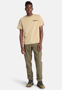 Camiseta de algodón beige con un pequeño logo impreso, combinada con pantalones cargo verdes oliva, que cuentan con bolsillos laterales y un ajuste relajado.