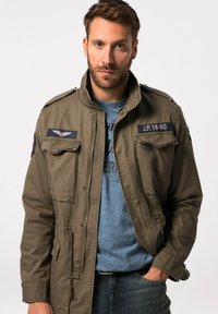 Giacca militare verde oliva con zip frontale, due tasche sul petto, epaulette sulle spalle e patch di insegna blu sulle maniche.