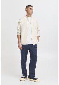 Cămașă bej deschis cu nasturi, tricou alb dedesubt, pantaloni bleumarin, adidași negri cu tălpi albe; croială lejeră, design minimalist.