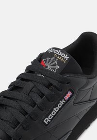 Svart Reebok Classic sneaker med overdel i läder, rund tå, texturerade detaljer och svarta snören; har logotyp och brittisk flagga på tungan.