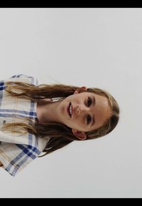 Niña joven con cabello largo y castaño, vistiendo una camisa a cuadros blanca y azul sobre un fondo claro y sencillo, sonriendo ligeramente.