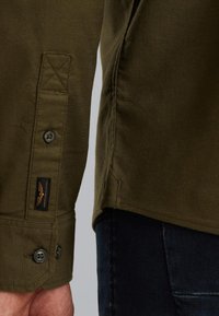 Camicia verde oliva con finitura testurizzata, dotata di polsini con bottone, dettagli cuciti e un'etichetta in tessuto sul lato.