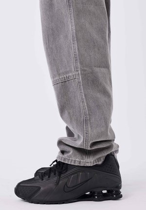Personne portant un jean gris ample et des baskets Nike Shox noires, debout sur un fond gris.