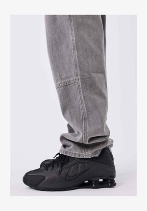 Persona che indossa jeans grigi larghi e sneakers nere Nike Shox, in piedi su uno sfondo grigio.