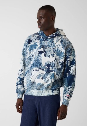 Man die een blauw-witte hoodie met bloemenpatroon en tekst "GUESS 1981" draagt, gecombineerd met donkerblauwe broek, staand tegen een effen achtergrond.
