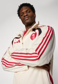 L.F.C. windbreaker jacket in cream with red and black accents. Features three red stripes on sleeves, zip closure, and elastic cuffs.