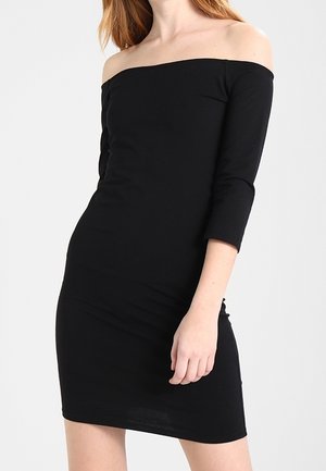 Kobieta w dopasowanej, czarnej sukience z odkrytymi ramionami i rękawami do łokcia, na jednolitym jasnym tle.