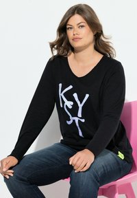 Schwarzes Langarmoberteil mit V-Ausschnitt, das mit weißen Buchstaben "KYST" versehen ist. Weicher Stoff, abgerundeter Saum, kombiniert mit dunkelblauen Jeans und grünem Logo-Akzent.
