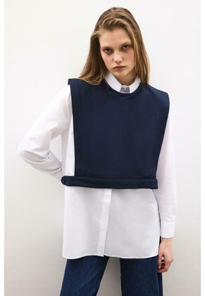 Młoda kobieta ubrana w białą koszulę na guziki pod bezrękawnikiem w kolorze granatowym, w połączeniu z ciemnoniebieskimi dżinsami, stojąca na tle białej ściany.